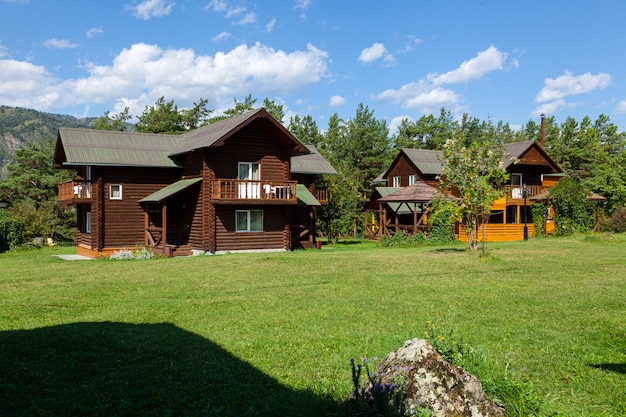 Two dark wooden log cabins sit on a vast, sunny green lawn, surrounded by a dense forest of pine trees under a bright blue sky with light clouds